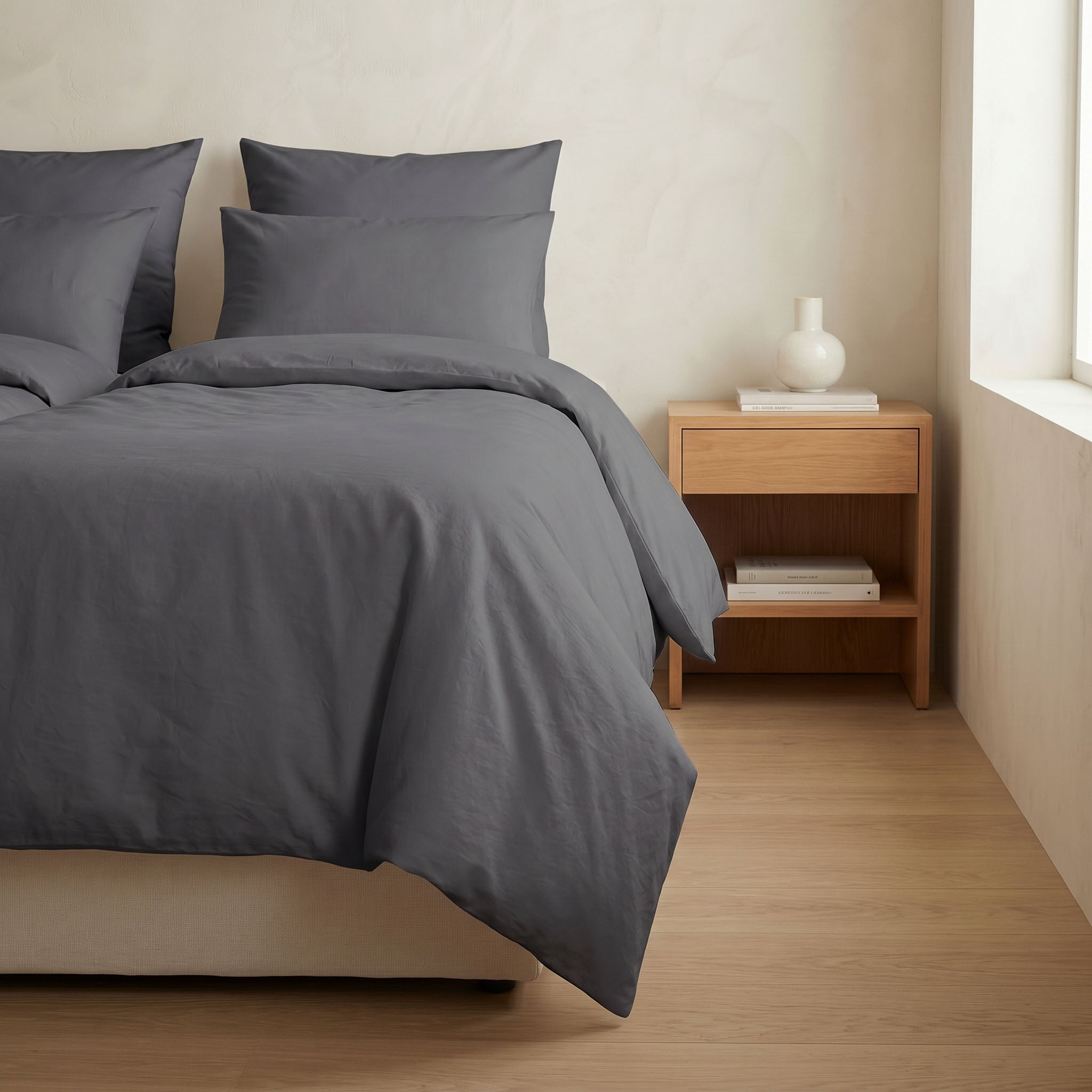 Plumelle wasbaar dekbed in Charcoal netjes opgemaakt op bed in lichte minimalistische slaapkamer met houten nachtkastje, naadloos 100% katoen